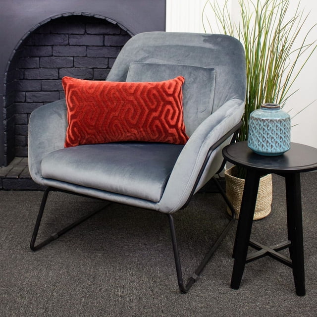 Zulu Luxury Feather Filled Cushion in red with a geometric pattern placed on a grey velvet armchair with black metal legs, beside a black round side table holding a blue ceramic vase, set in a room with a black brick fireplace and tall green plants in a woven basket, suitable for decorative use and added comfort.