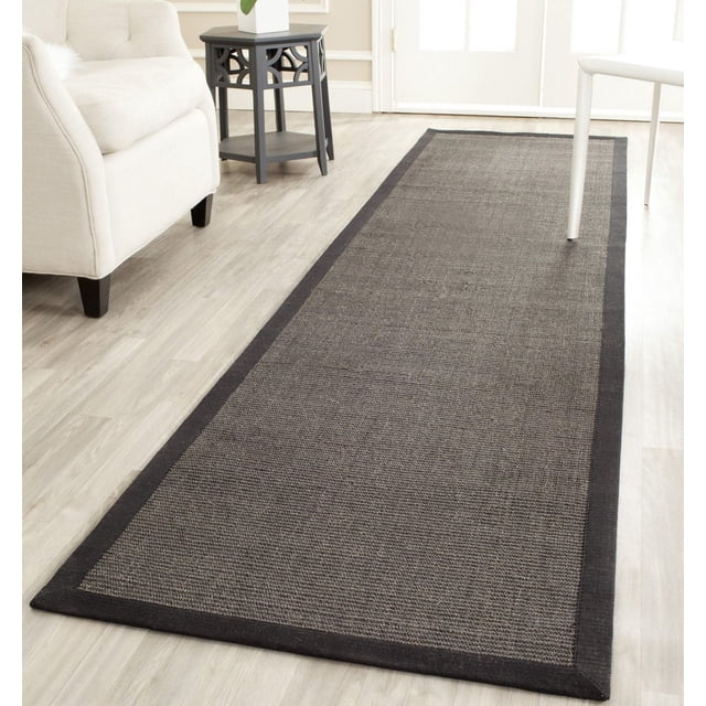 Safavieh Natural Fiber Forrester Border Area Rug with dark grey woven sisal texture and black border, laid on light wood flooring in a bright room with beige armchair, black side table, and white chair, ideal for coastal or casual interiors.