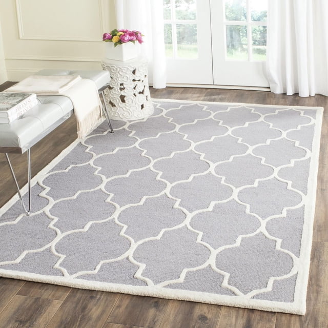 Safavieh Cambridge Kevin Geometric Area Rug in light grey wool with a white geometric trellis pattern, hand-tufted with plush cut and high/low loop pile, displayed on a hardwood floor near a white carved stool and a bench with books and a throw, ideal for modern home or office decor.