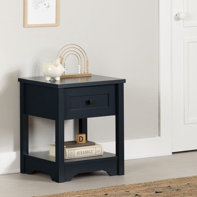 South Shore Hazen Nightstand for Child in navy blue with shaker-style drawer and minimalist black knob, finished on all sides with a bottom shelf holding a Scrabble book and wooden block, topped with a ceramic piggy bank and wooden bead maze, placed in a child's room with light walls and white door, suitable also as a side table for reading spaces.