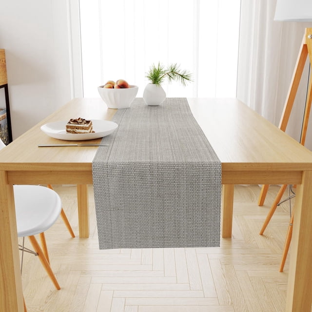 Fabstyles Casual Classic Cotton Table Runner in neutral gray woven cotton fabric measuring 13x54 inches, laid on a light wood dining table with a white plate holding a slice of layered cake and fork, a white bowl with peaches, and a small white vase with pine sprigs, in a bright room with white curtains and light wood flooring, also suitable as a kitchen or patio table runner.