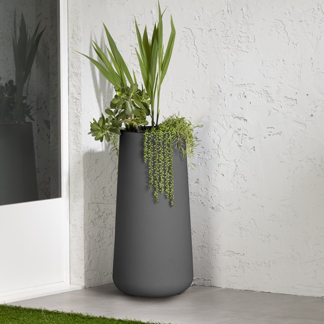 South Shore Dalya Tall Outdoor Pot Planter in dark gray with a matte finish, made of weather-resistant polyethylene resin, featuring a large tapered cylindrical shape holding up to 10 liters of soil, filled with assorted green plants including tall palm leaves and trailing succulents, placed on a concrete floor against a textured white wall near a glass door, suitable for outdoor garden or patio decoration.