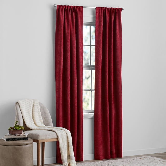 Mainstays Sheen Rod Pocket Blackout Window Curtains Panel Pair in deep red, 37" x 84", hanging on a silver rod over a window with natural light. The blackout panels have a subtle sheen texture that blocks light and reduces noise. A beige chair with a cream throw and a small ottoman with books and a plant are beside the window in a minimalist room with light gray walls and wood flooring, suitable for enhancing privacy and temperature control.