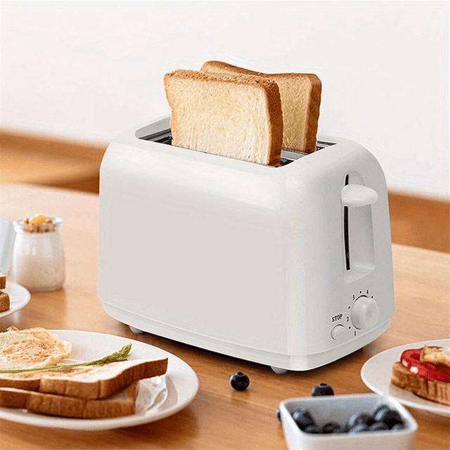 Kitchen Fully Automatic Breakfast Machine Toaster Bread Toaster Home Breakfast Toaster Heater Tool US Plug in white. The toaster features widened double slots for accommodating bread of various thicknesses, with automatic centering to ensure even toasting. It includes a pull-out crumb tray for easy cleaning and operates on AC110V with a US plug. The image shows the toaster on a wooden table with two slices of bread being toasted, surrounded by breakfast items like toast, berries, and spreads.