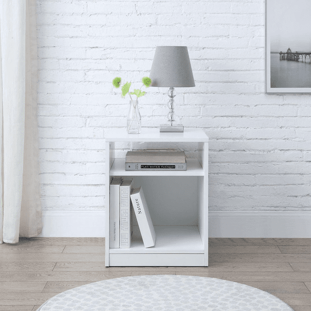 Mainstays Open Shelf Nightstand in white laminated particle board with a rectangular shape, featuring a top surface holding a clear glass vase with green flowers and a grey fabric table lamp, an open middle shelf with a closed book, and a large lower cubby storing three books, set on a light wood floor against a white brick wall with a round patterned rug in front. This nightstand is suitable for storing bedside essentials within easy reach.