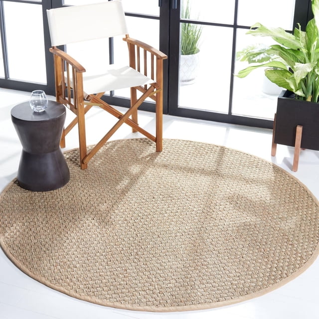 Safavieh Tapis à Bordure en Tonnelle en Fibre Naturelle, Rond avec texture Tissée sisal beige Naturel, Placé dans une Pièce Lumineuse avec des Planchers Blancs. Positionné sous une Chaise en Toile Blanche à Cadre en Bois et à Côté d'Une table d'Appoint Sablier Noire avec un Verre d'Eau. un Cache-Pot Noir avec des Plantes Vertes Se Tient Près de Grands Panneaux de Fenêtre. Convient aux Intérieurs style Maison de Plage Côtière Ou Décontractée.