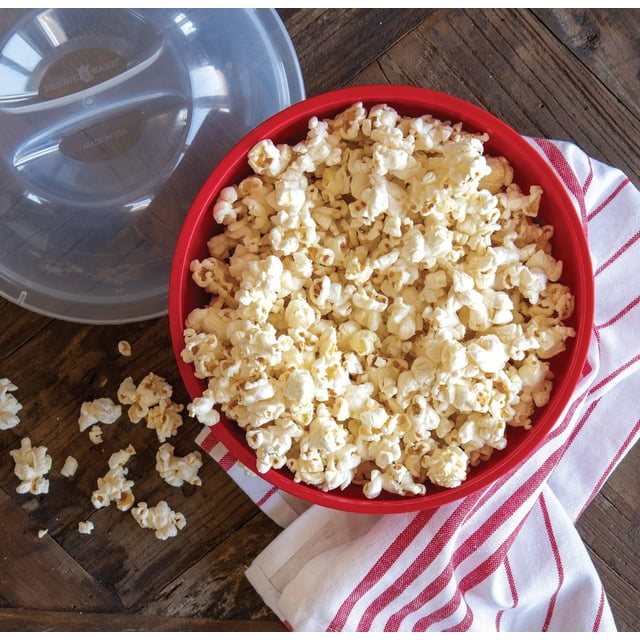 Nordic Ware Quick Pop Single Serve Popper with red BPA-free plastic bowl filled with air-popped popcorn, clear lid beside it showing brand markings, placed on a wooden table over a white and red striped kitchen towel with some popcorn scattered nearby.
