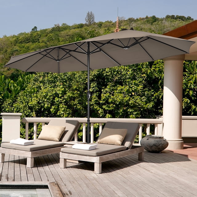 Costway Parasol de Patio Double Face de 15 Pi, Couleur Café, avec Auvent de 15 Pi x 9 Pi, Doté d'Un Cadre Métallique Robuste et d'Un Mécanisme à Manivelle. le Parasol Est Installé sur une base Métallique Croisée avec Deux Sacs de Sable pour la Stabilité, Idéal pour une Utilisation Extérieure sur les patios, les Jardins Ou les Zones au Bord de la Piscine. l'Image Montre le Parasol Offrant de l'Ombre sur Deux Chaises Longues sur une Terrasse en Bois Entourée de Verdure.