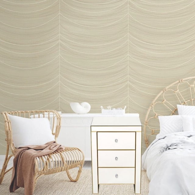 Nightstand with three drawers and clear knobs in a gold mirrored finish, serving as a bedside storage cabinet between a rattan chair with white cushion and a bed with white bedding, set against a beige textured wall in a bedroom.