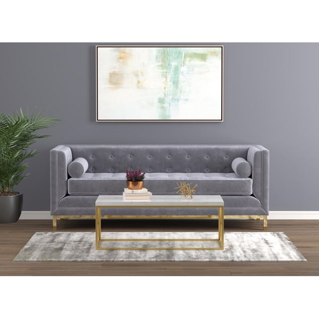 Safdie & Co. Coffee Table 44L Marble Gold Metal with a rectangular white marble top and gold metal frame, placed before a grey velvet tufted sofa with bolster pillows on a light grey rug over wood flooring, complemented by a potted plant and abstract wall art, suitable for modern living room settings.