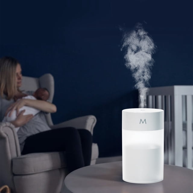Mini Air Purifier Humidifier with Colorful Night Light, featuring ultrasonic cool mist and essential oil compatibility, placed on a table in a dimly lit room. The compact white cylindrical design emits visible mist, ideal for bedroom or nursery use. A woman holding a baby is seated in the background. Includes adjustable mist output, color-changing LED lights, silent operation, and auto shutoff functionality.