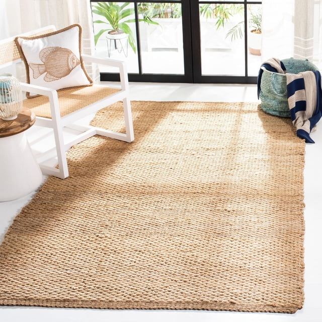 Safavieh Natural Fiber Bryon Braided Area Rug in natural beige braided sisal on a white floor, shown in a bright room with a white chair with woven seat and fish-patterned pillow, a wooden side table, and a blue woven basket with a striped blanket, suitable for coastal or casual beach house style interiors.