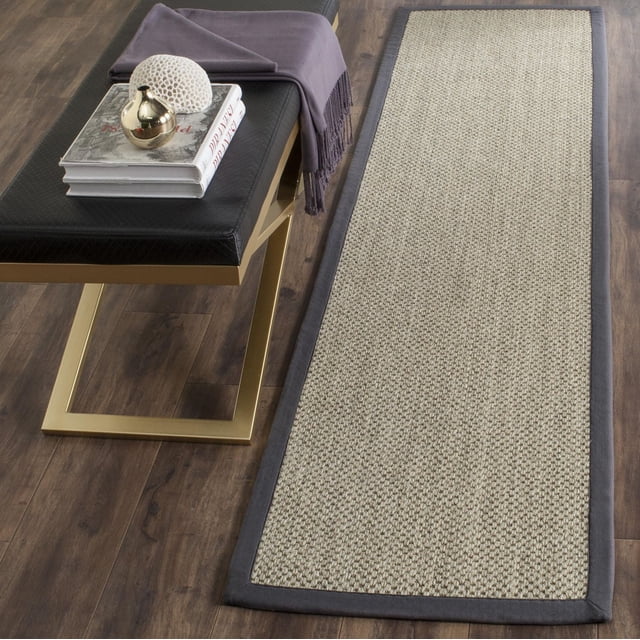 Safavieh Tapis à Bordure de Genévrier en Fibre Naturelle dans une Vue Inclinée du Haut vers le Bas sur Plancher de Bois Franc Foncé, Présentant une texture Tissée en sisal Naturel avec une Bordure en Tissu Gris Foncé, Placé à Côté d'Un Banc Moderne avec des Pattes en Métal Doré et un Siège Coussiné Noir, style avec des Livres, une Pièce de Déco Corail Blanc, et un Petit vase Doré, Convient pour un style Côtier Ou Décontracté de Maison de Plage et comme Couloir Coureur.