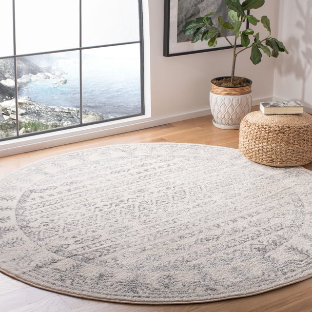 SAFAVIEH Tulum Flint Distressed Southwestern Area Rug, round with soft grey and off-white rustic geometric patterns, placed on light hardwood flooring near a large window with ocean view, beside a woven pouf with a book and a potted plant in a white decorative planter, ideal for enhancing room décor with warm, inviting character.