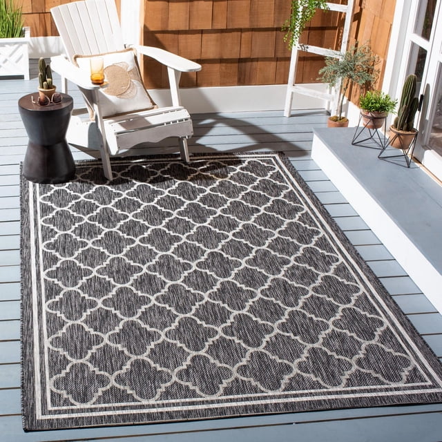 SAFAVIEH Courtyard Claudia Tapis Géométrique Intérieur/extérieur sur une Terrasse en Bois Gris Pâle, avec une base en Polypropylène Gris Foncé avec un Motif Géométrique Trèfle Blanc Cassé et des Bords Bordés, Style avec une Chaise Adirondack Blanche, table d'Appoint Noire avec cactus et Lunettes de Soleil, et Plantes en Pot par un Mur en Bardeaux de Bois et une Porte en Verre, Idéal pour une Utilisation durable Intérieure et Extérieure.