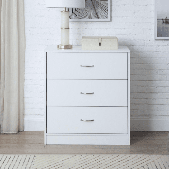 Mainstays 3-Drawer Dresser in white finish with three metal-handled drawers, front-facing against a white brick wall and beige curtain, topped with a white and gold lamp and beige storage box, on light wood flooring with a patterned rug, suitable as kids' furniture or compact storage.