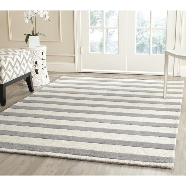 SAFAVIEH Cambridge Keaton Striped Wool Area Rug featuring hand-tufted wool in soft grey and cream horizontal stripes, displayed on light wood flooring in a bright room with a chevron-patterned bench and white ceramic side table with orchid, suitable for modern home or office decor.