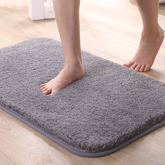 Chmadoxn Bathroom Rugs, a thick gray bath mat made of polyester with a soft, high-density fiber surface and a non-slip Thermo Plastic Rubber bottom. Measuring 40x60cm (15.75x23.62 inches), this absorbent mat is suitable for bathrooms, kitchens, and indoor doorways. The image shows a foot pressing into the plush surface, highlighting its softness and comfort.