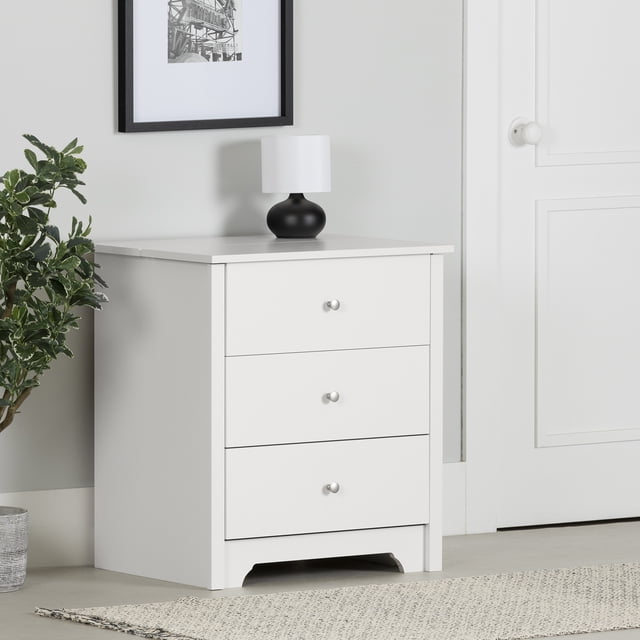 South Shore Vito Nightstand with Charging Station and Drawers for Adult in pure white, front-facing with three smooth-gliding drawers with metal knobs, a flat top holding a black lamp with white shade, featuring a sliding tray charging station with cord management, set against a light grey wall near a white door and green plant, suitable for contemporary bedroom storage and device charging.