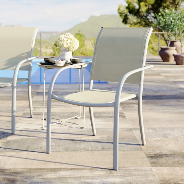 Mainstays Steel Outdoor Stacking Chair in greige with rust-resistant powder-coated steel frame and UV-resistant sling fabric, shown on a tiled patio beside a small round table with a white flower vase and black cups, suitable for outdoor patio seating and stackable for easy storage.