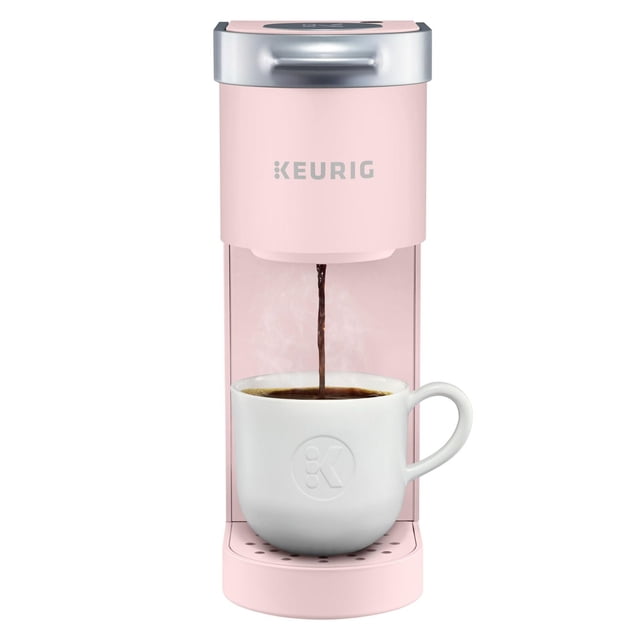 Keurig K-Mini Single Serve K-Cup Pod Coffee Maker in pink with silver top pouring steaming coffee into a white ceramic mug embossed with a 'K' logo, compact design ideal for small spaces and quick single-cup brewing.