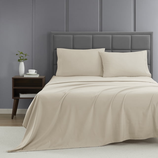 Nestl Cal King Sheet Set, 1800 Series Microfiber Deep Pocket 4 Piece Bed Sheet Set displayed on a neatly made bed. The set includes a fitted sheet, flat sheet, and two pillowcases in a soft beige color. The bed is styled with a grey upholstered headboard and a wooden nightstand holding a white vase and cup. The microfiber fabric is double-brushed for softness, resistant to pilling, and features deep pockets for mattresses up to 16 inches. The setting is a modern bedroom with a grey panelled wall and a light carpeted floor.