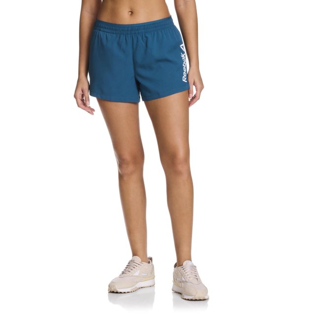 Reebok Women's Staple Running Short with Mesh Lining in blue, front view on a model from waist to mid-thigh, featuring elastic waistband with drawcord, quick-drying lightweight polyester fabric, built-in mesh liner for breathability, and white Reebok Delta logo on the left leg, suitable for running, training, or casual wear.