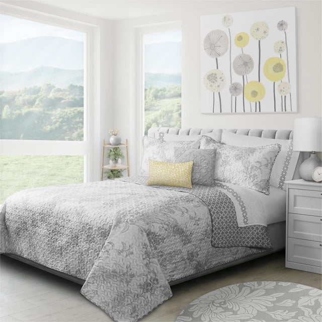 Safdie & Co. Quilt 3PC Set K Transitions Alloy in grey polyester microfiber on a bed with matching shams and a yellow accent pillow in a bright bedroom with large windows showing green hills. The quilt features a floral and swirl pattern, designed for comfort and durability. The set adds a subtle elegant touch to bedroom decor.