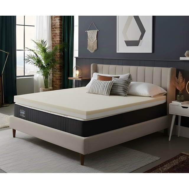 Millano Memory Foam Topper-Double on a beige upholstered bed frame with vertical channel tufting in a modern bedroom. The 4-inch topper combines two inches of supportive foam and two inches of antimicrobial COPPERGEL memory foam for cooling and pressure relief. The room features decorative pillows, a nightstand with a lamp, a green plant by a large window with dark green curtains, and geometric wall art.