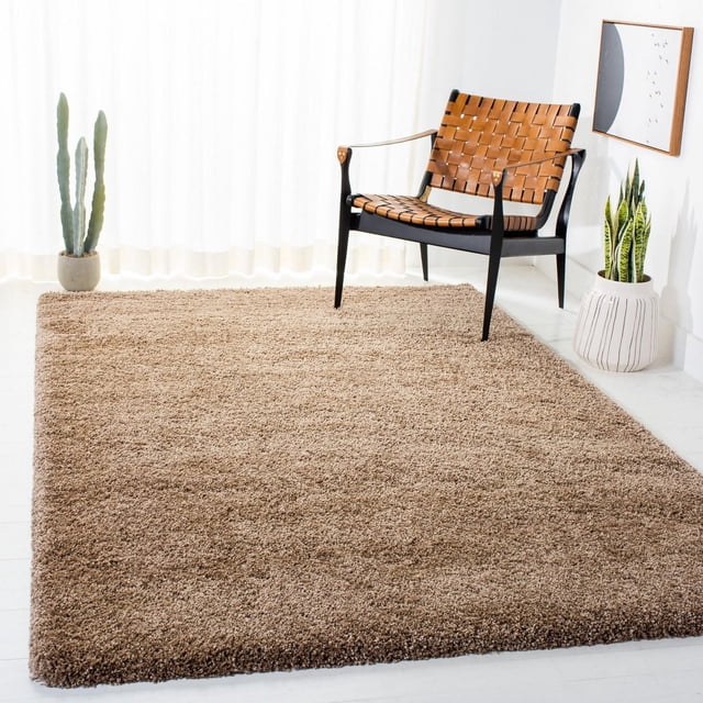 Safavieh Milan Harlow Solid Shag Area Rug in beige, placed in a bright room with white floors and sheer curtains. The plush synthetic yarn rug is under a black-framed chair with woven tan leather straps, flanked by a cactus and a snake plant in white pots. This durable accent rug adds soft texture and warmth to contemporary spaces and is often used to define seating areas.
