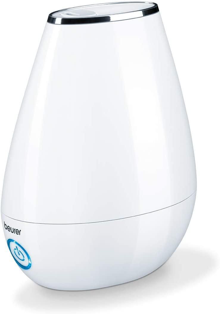 Beurer Humidifier front-facing tabletop view of a compact white humidifier with removable translucent water tank, top mist outlet and control buttons. Shows ultrasound humidification technology, night mode, two output levels, white/blue/red LED indicators and automatic shut-off. Includes 15 aroma pads and cleaning brush; often used to add a gentle scent to small offices or bedside tables.